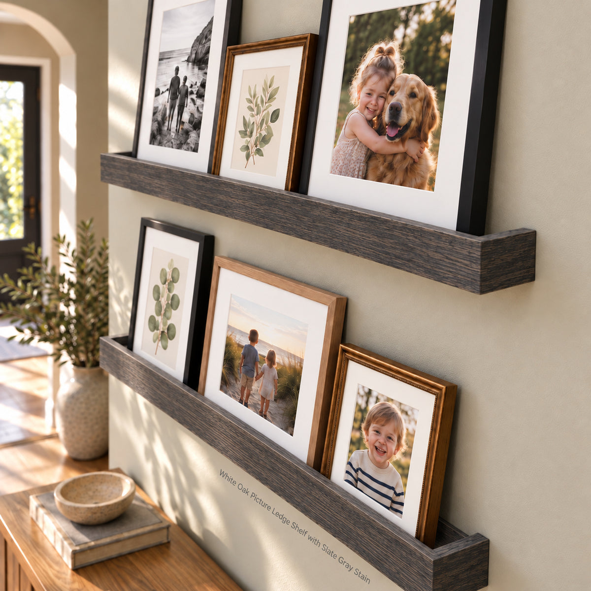 Two dark gray picture ledge shelves mounted on a beige wall, styled with multiple framed family and botanical photographs in black and natural wood frames. The scene is set in a warm, sunlit room with a wooden floor and a potted plant in the background.